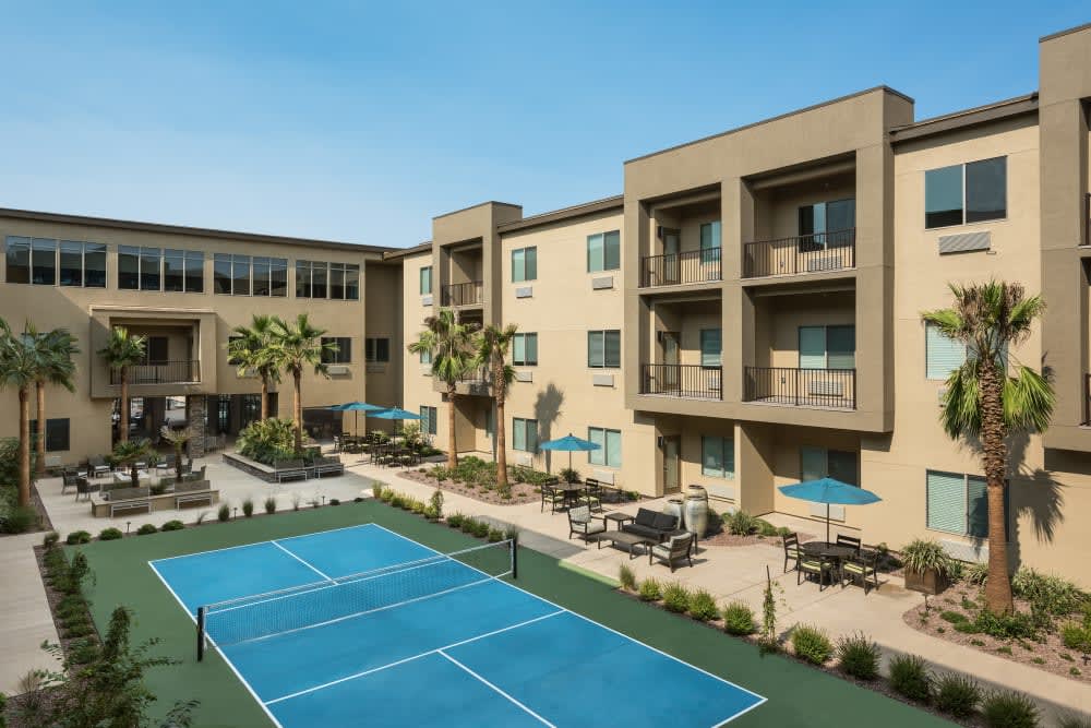A modern apartment complex featuring a tennis court and outdoor seating area, surrounded by palm trees.