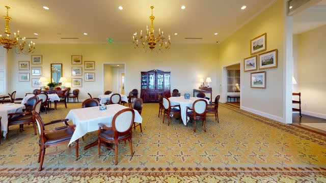 A spacious dining room with elegant classic furniture and a central chandelier, featuring tables set with white tablecloths.