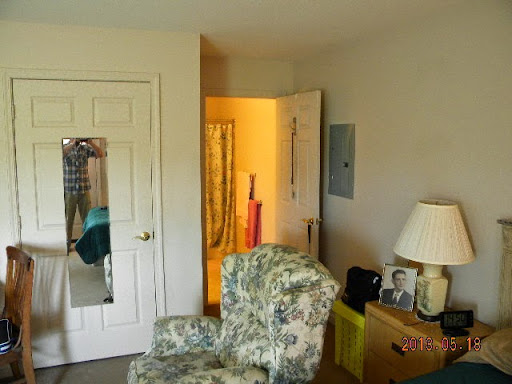 A cozy bedroom featuring a patterned chair, lamp, mirror, and an open door leading to another area.