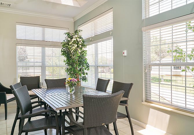 A bright dining room with a large table, surrounded by chairs and illuminated by natural light streaming through multiple windows with blinds.