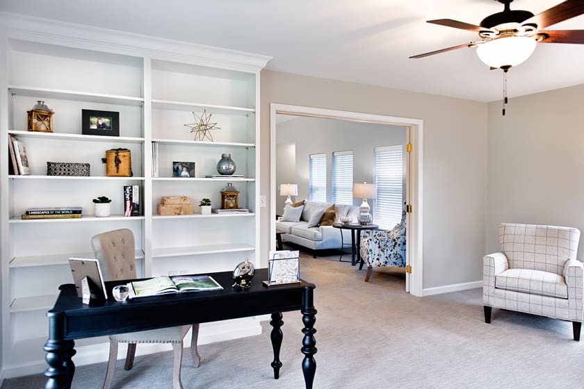 A spacious home office with a black desk and built-in shelves, leading into a cozy living room filled with natural light.