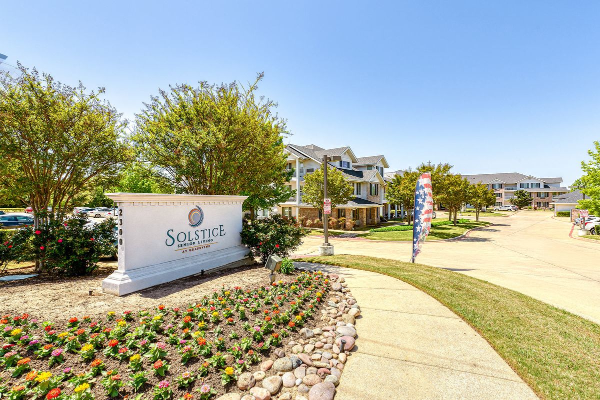 A well-landscaped entrance to a suburban residential community with a prominent sign and houses in the background.