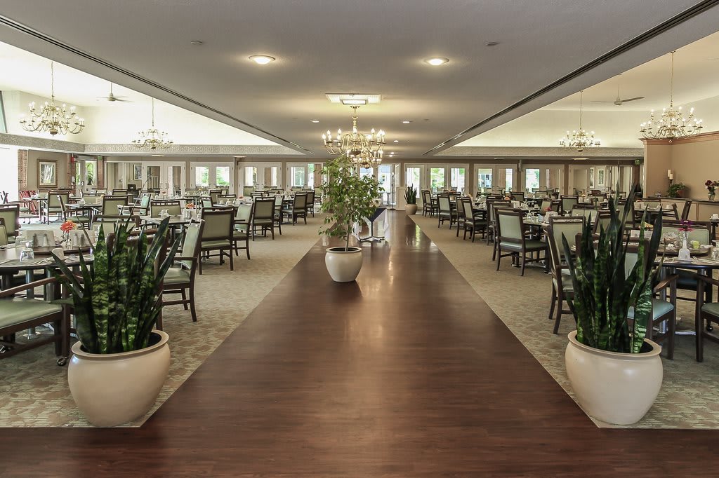 A spacious dining hall with elegant chandeliers, neatly arranged tables and chairs, and decorative plants.