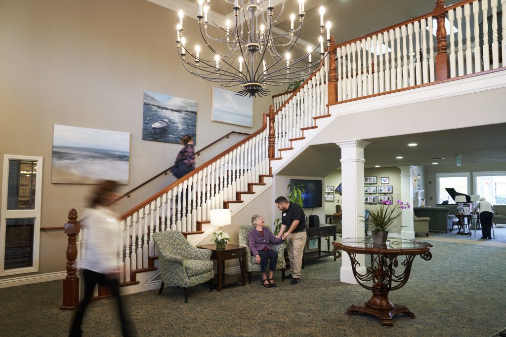 A spacious interior of a nursing home with a grand staircase, chandelier, and seating area where an assistant is helping a resident.