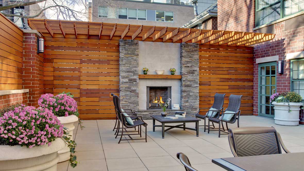 A cozy outdoor patio with a fireplace and comfortable seating under a wooden pergola in an urban setting.
