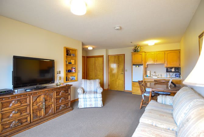 A cozy living space featuring a television, armchair, bookshelf, kitchenette, and dining area.
