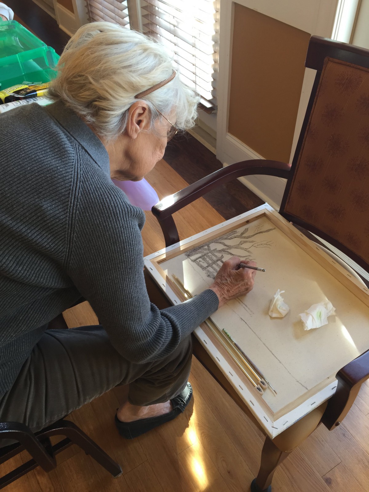 An elderly woman engaged in painting at a table, focused on her artwork.