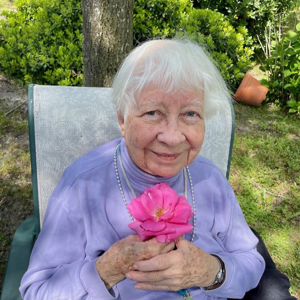 An elderly woman sitting outdoors in a garden, holding a pink flower.