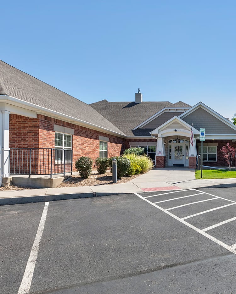 An exterior view of a building with a parking lot, featuring a brick facade and peaked roof.