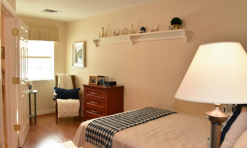 A cozy and minimalist bedroom featuring a neatly made bed, a wooden dresser, a chair by the window, and decorative items on a wall shelf.