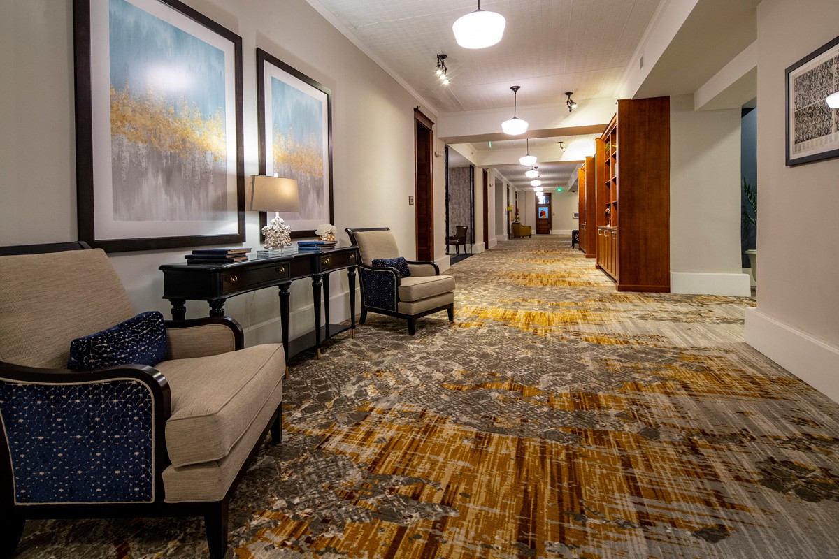 A well-lit hotel hallway with decorative artwork, comfortable seating, and patterned carpet.