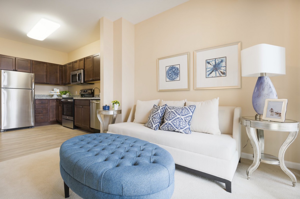 A modern living area with a comfortable white sofa and blue ottoman, seamlessly connected to a kitchen.