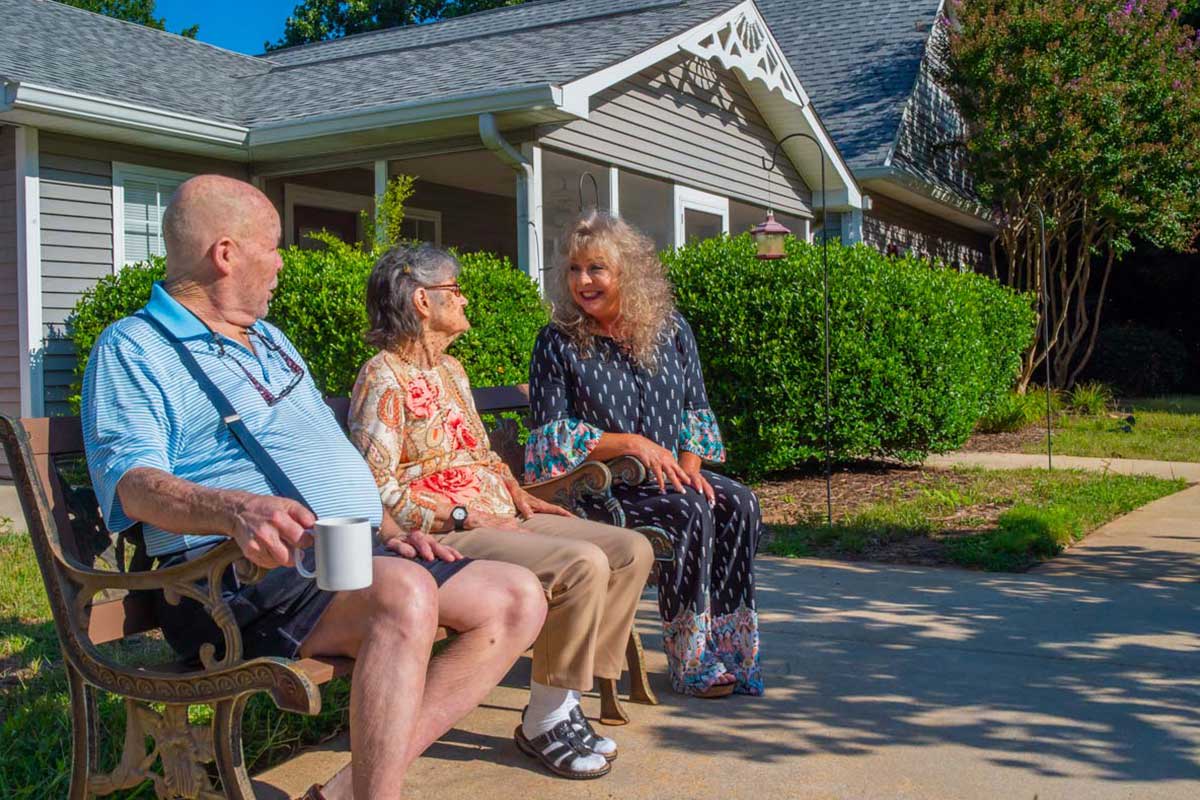 Three seniors sitting on a bench, enjoying a conversation outside a retirement community.