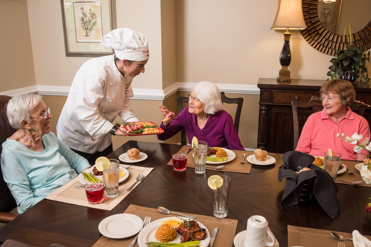 A chef serving a meal to elderly guests in a restaurant setting.