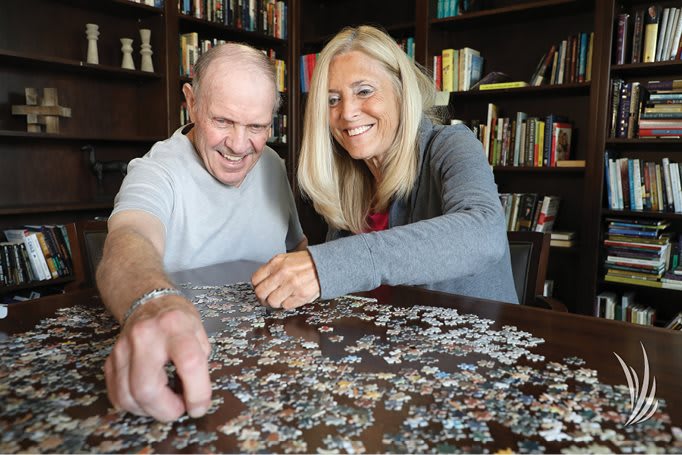 Two people happily working on a jigsaw puzzle together at a table in front of a bookshelf.