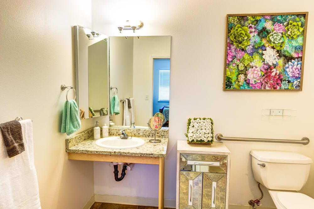A brightly lit bathroom featuring a sink with a mirror, towels, and colorful artwork on the wall.