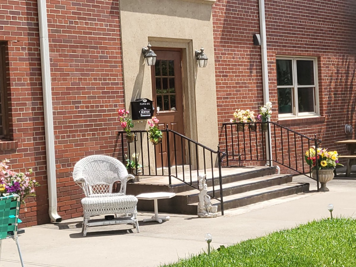 A quaint front porch with a wicker chair and flowers by the steps of a brick building.