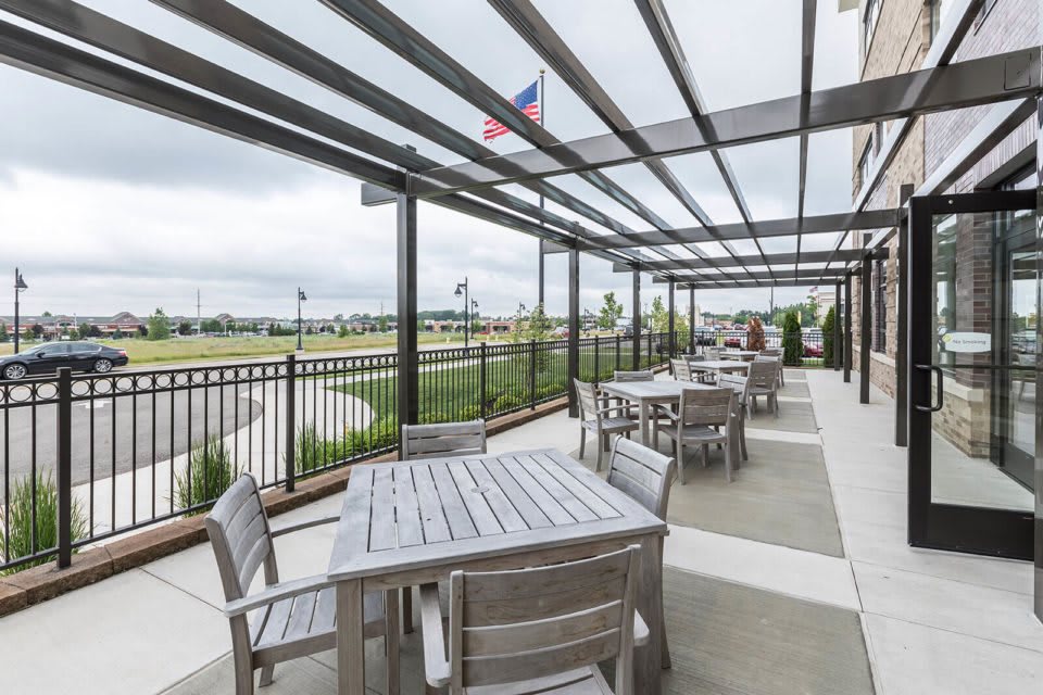 An outdoor patio area with tables and chairs under a pergola, overlooking a landscaped area with an American flag in the background.