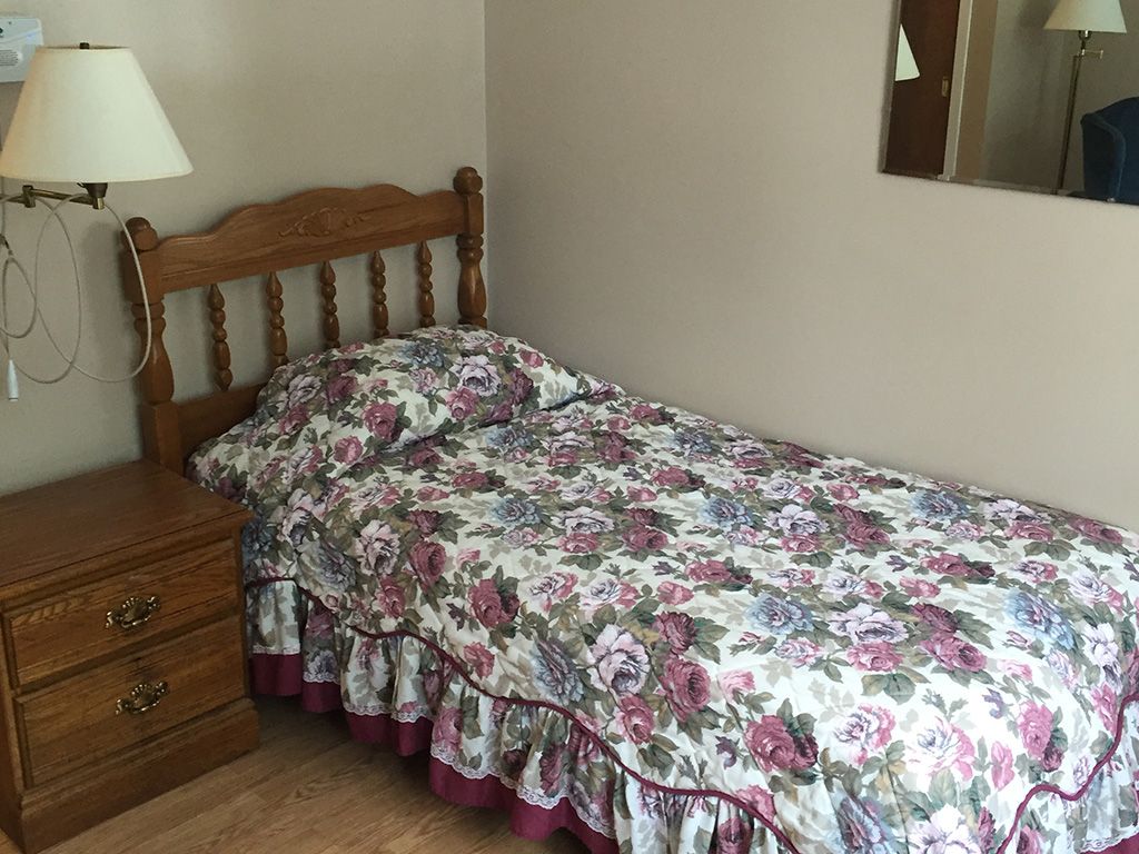 A tidy bedroom with a single bed covered in a floral bedspread, accompanied by a wooden nightstand and lamp.