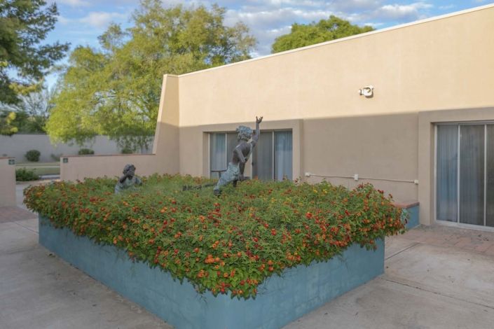 An outdoor sculpture in a garden setting, surrounded by greenery and adjacent to a modern building.