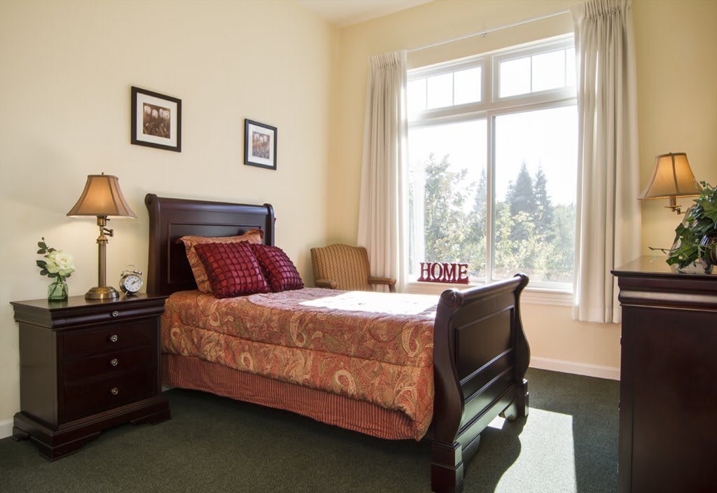 A cozy bedroom featuring a wooden bed with patterned bedding, a nightstand, and a large window flooding the room with natural light.