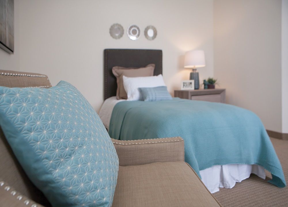 A cozy bedroom featuring a neatly made bed with blue bedding, a side table with a lamp, and decorative pillows.