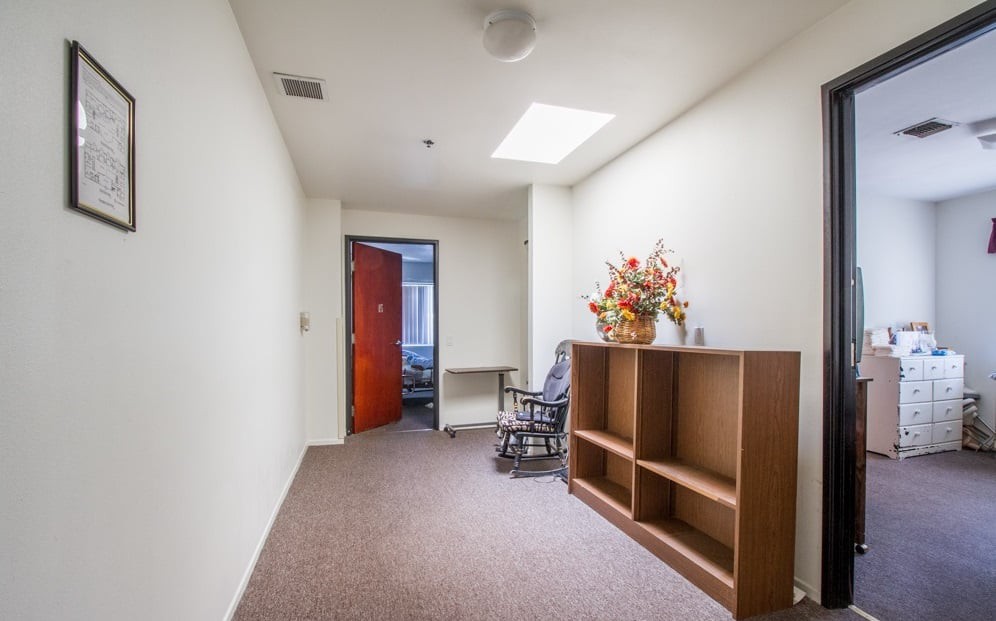 A narrow hallway with a rocking chair, shelving unit, and a flower arrangement near the doorway.