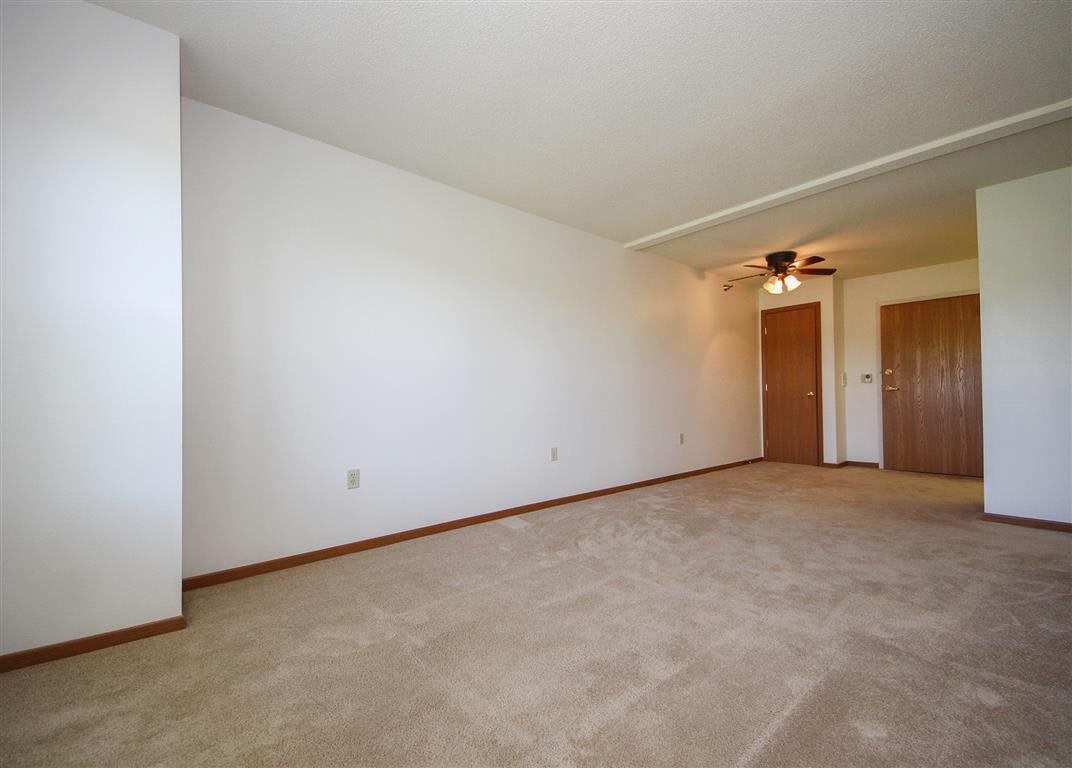 An empty room with a carpeted floor, white walls, and a ceiling fan near the doorway.