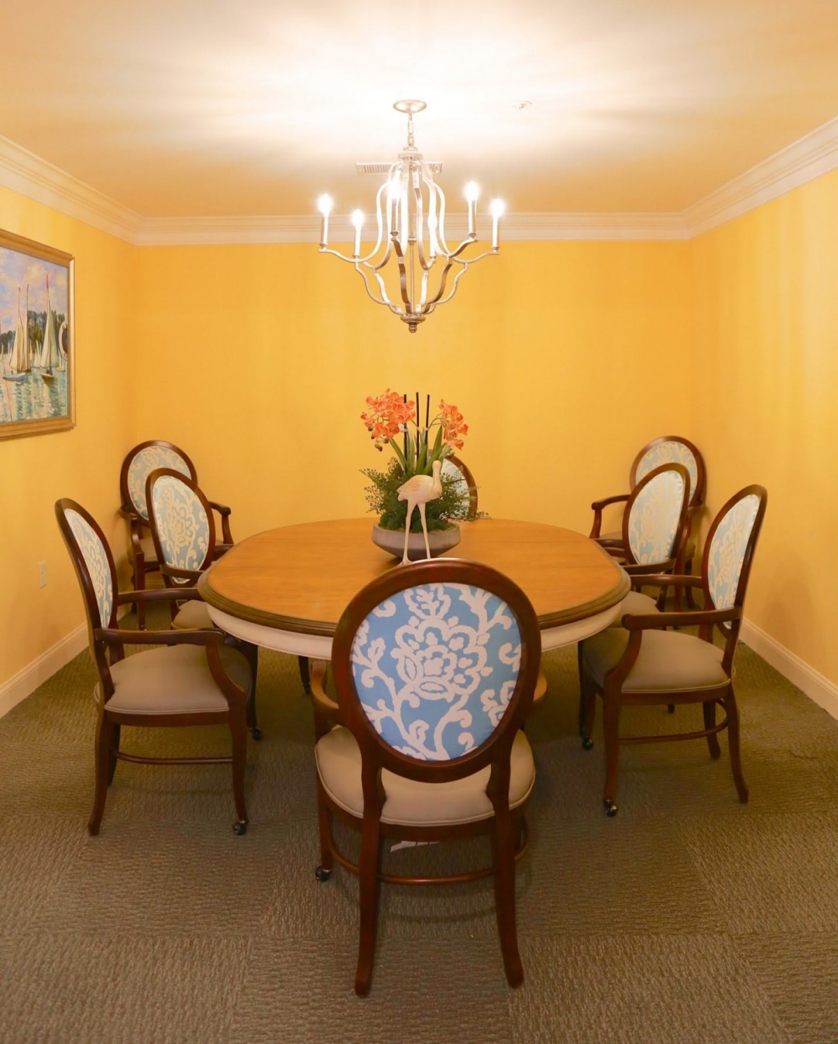 A dining room with a round table surrounded by six upholstered chairs, a chandelier overhead, and yellow walls.
