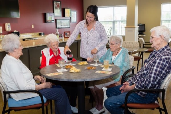 A caregiver serves drinks to a group of elderly people sitting around a table in a communal setting.