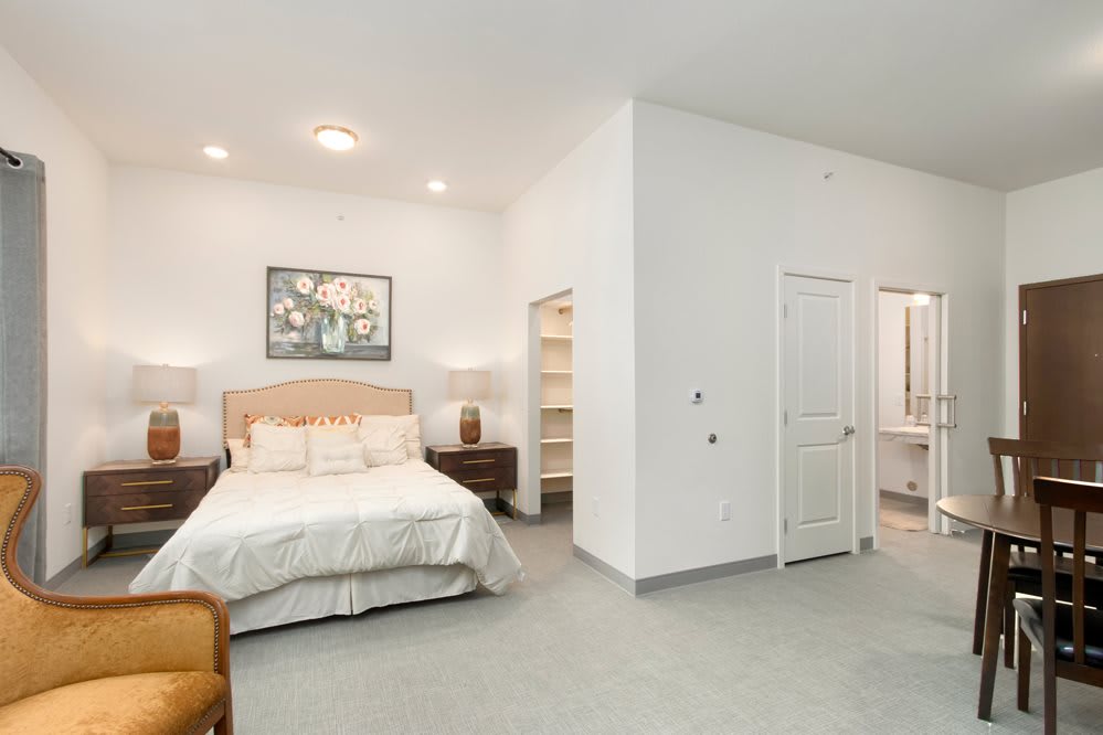 A modern, minimalist bedroom with white walls, a bed with neutral bedding, and carpeted flooring. The room features nightstands on either side of the bed and artwork hanging above.