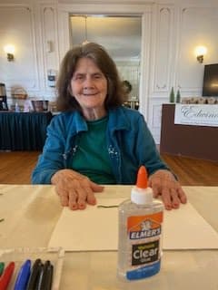 A woman sits at a table with a glue bottle, smiling while engaging in a crafting activity indoors.