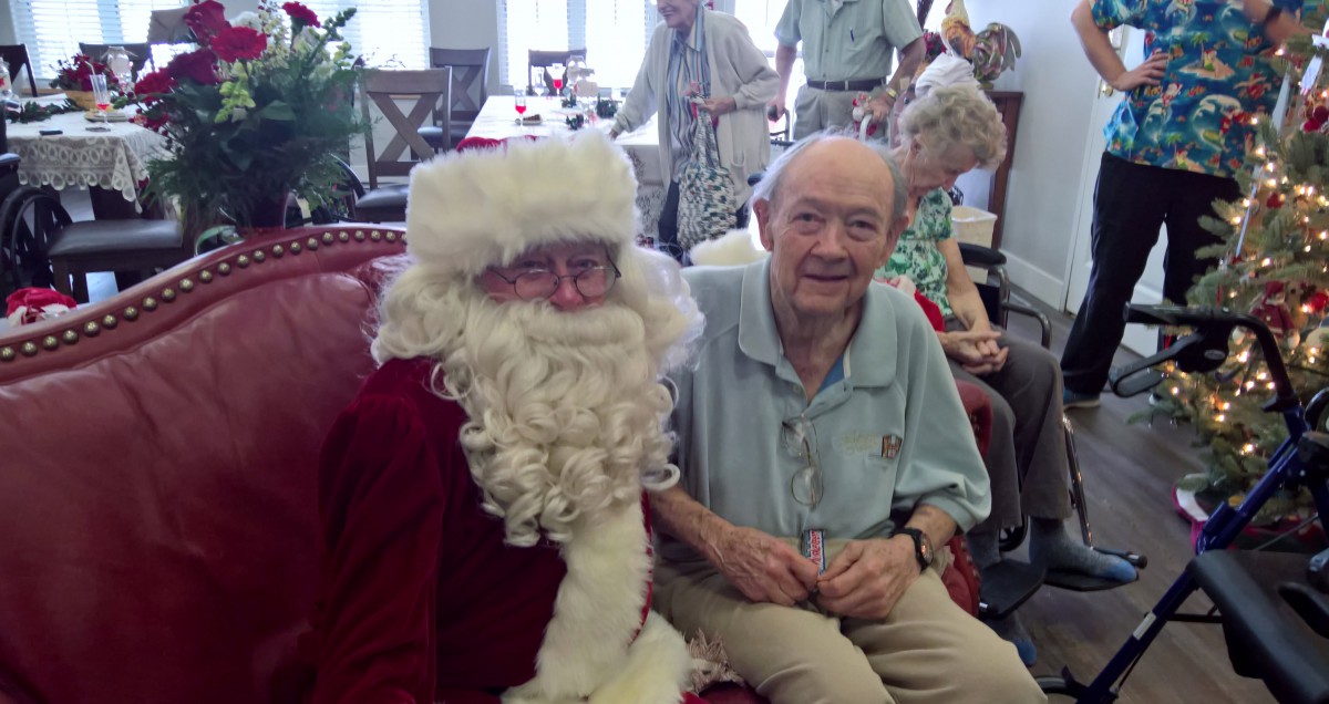 An elderly man sitting with someone dressed as Santa Claus during a holiday gathering.