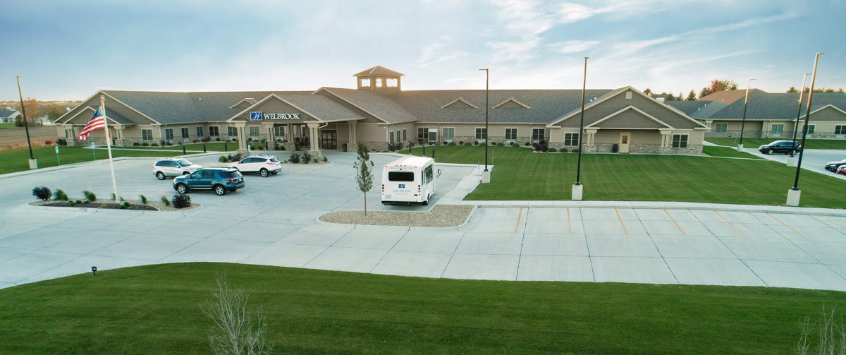 A building with a large parking lot and green lawn under a cloudy sky.