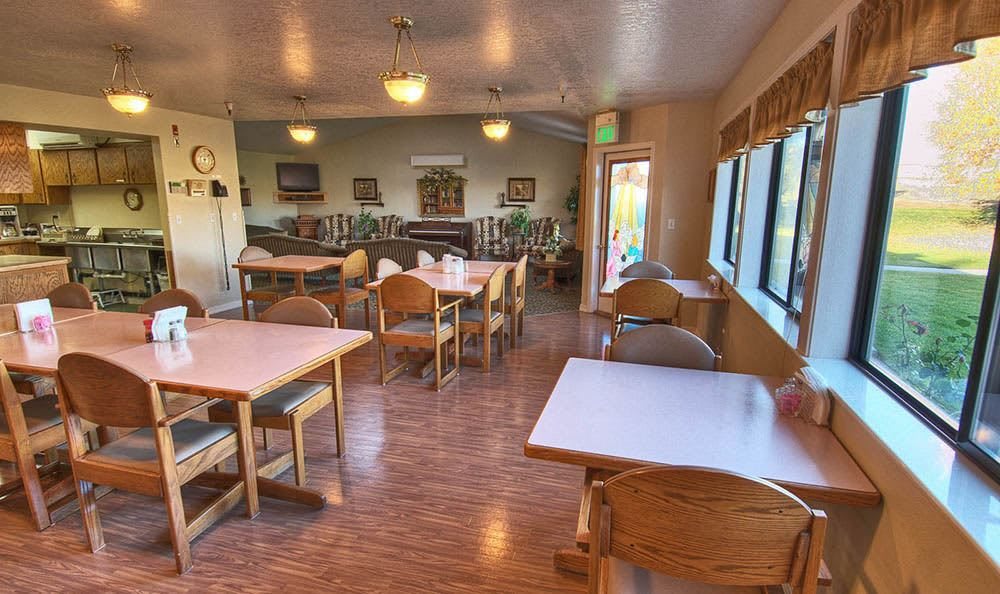 A cozy dining area with wooden tables and chairs, ambient lighting, and large windows providing natural light.