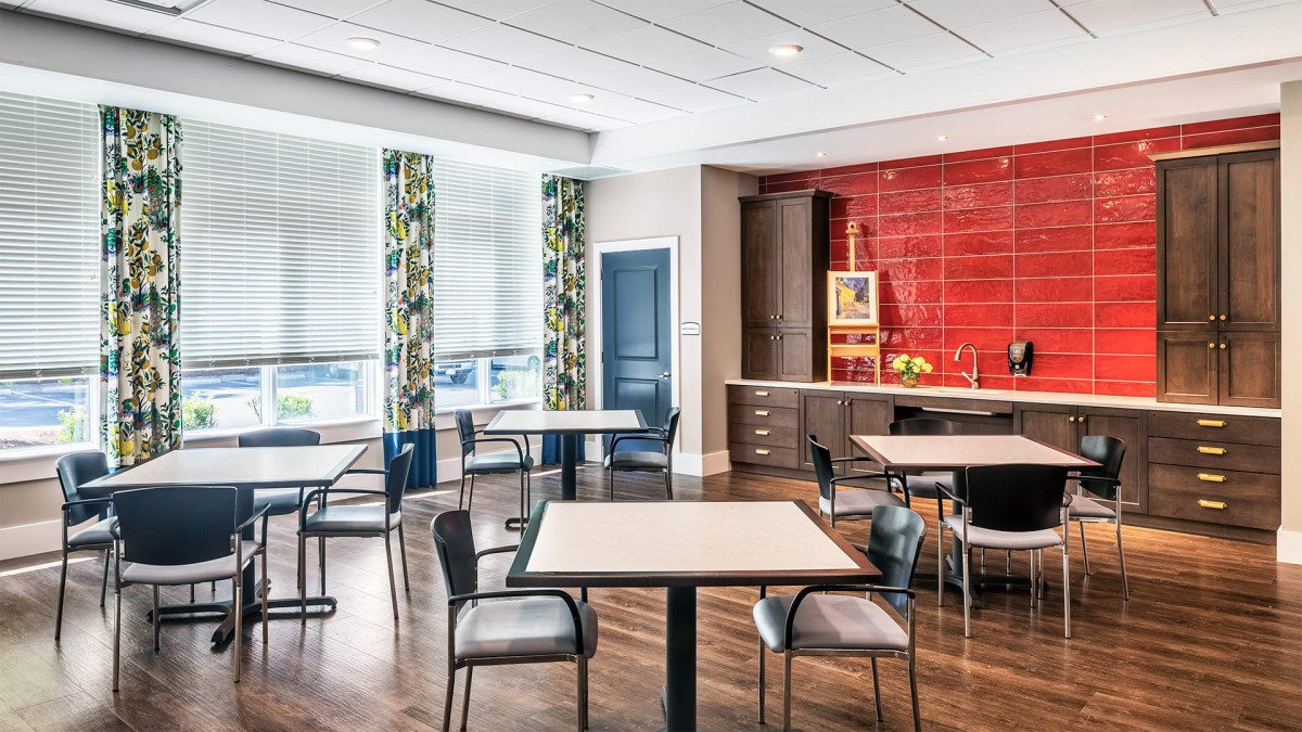 A modern indoor dining area with tables and chairs, featuring bright natural lighting and a vibrant red accent wall.