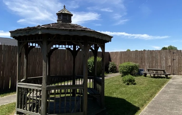 A wooden gazebo in a landscaped garden with surrounding bushes and a wooden fence under a blue sky.