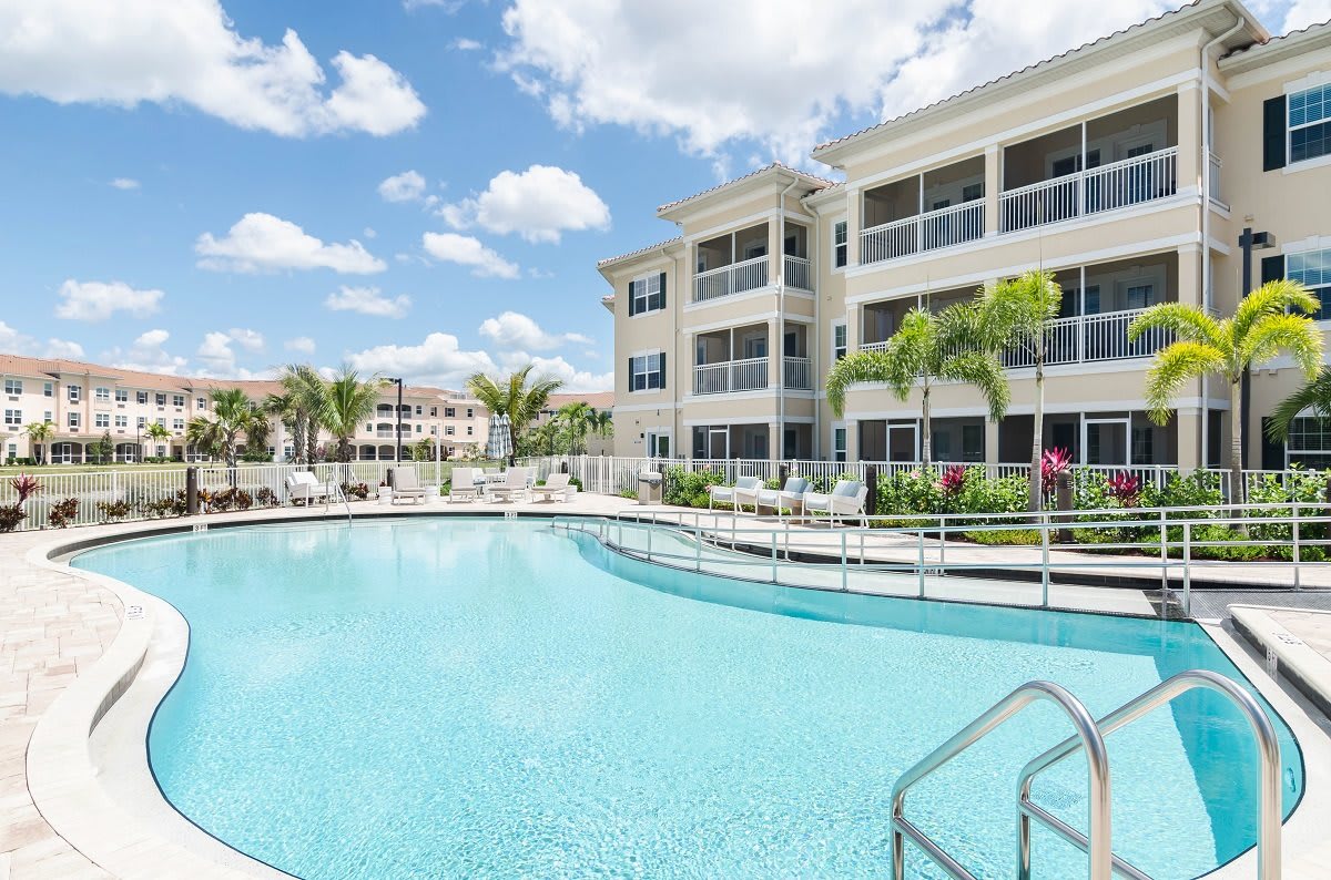 A modern apartment complex with a large swimming pool and surrounding palm trees under a partly cloudy sky.