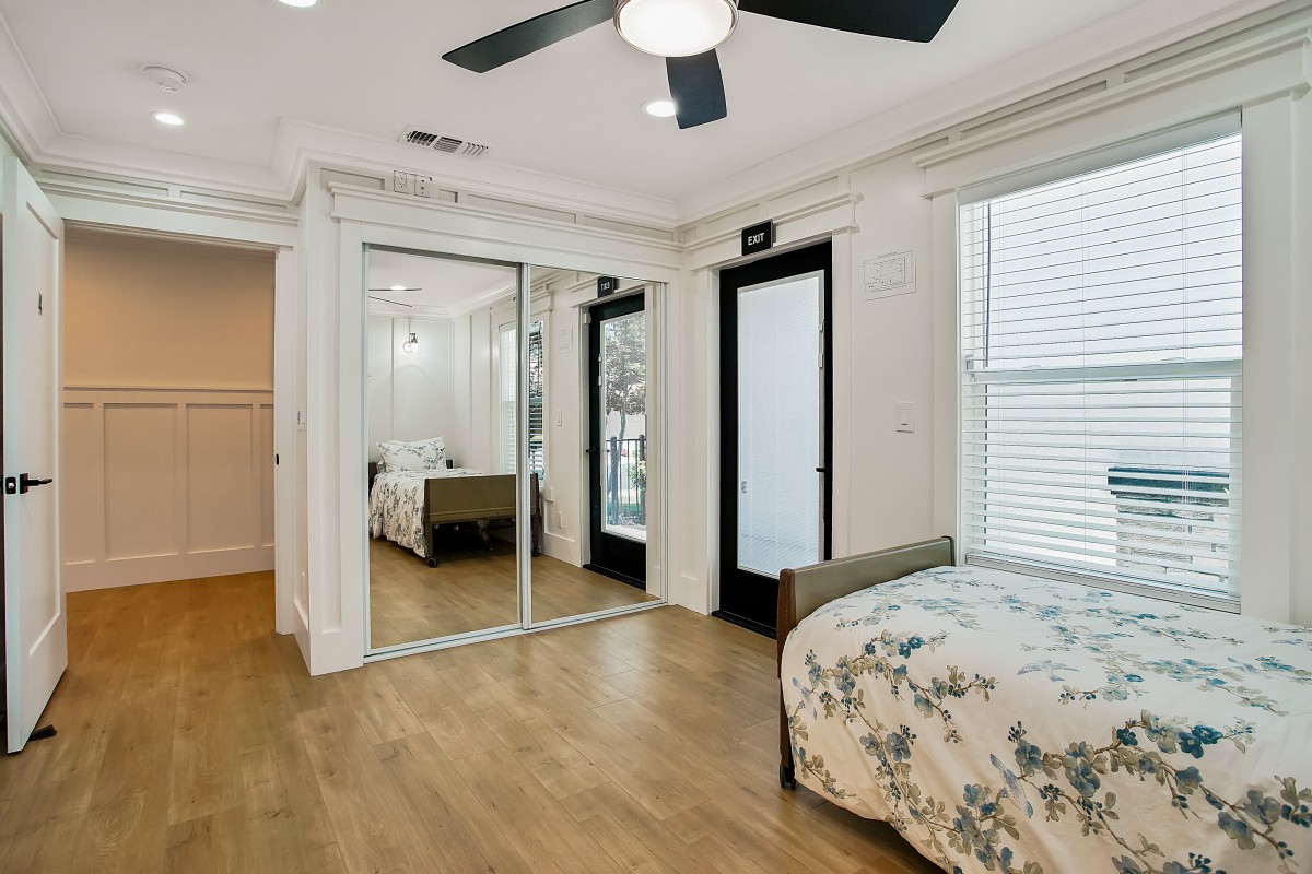 A modern bedroom featuring hardwood floors, a ceiling fan, mirrored closet doors, and a neatly made bed with floral bedding.