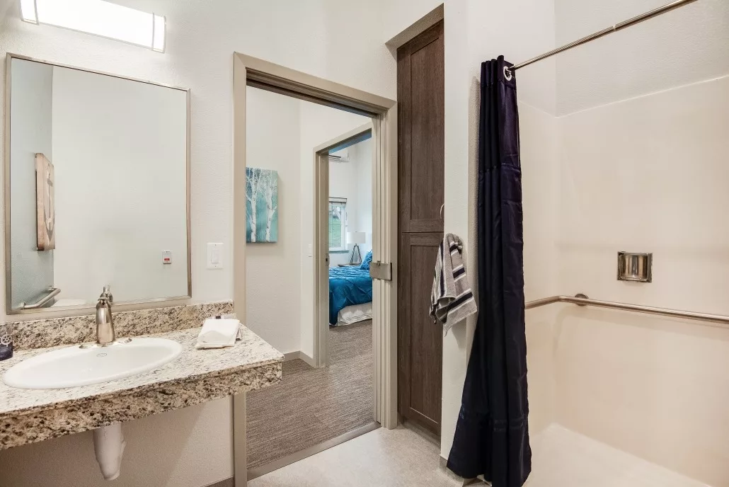 A modern bathroom with a shower, sink, and large mirror, featuring a view into an adjoining bedroom.
