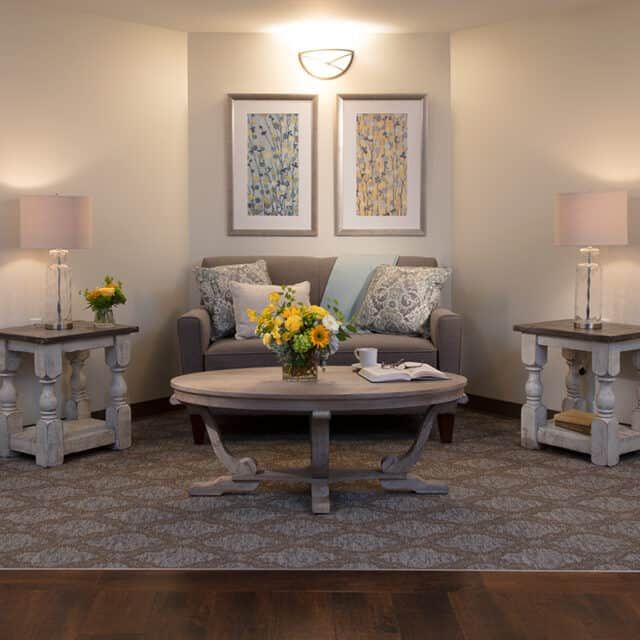 A cozy living room with a sofa, decorative pillows, and a central coffee table adorned with flowers, framed by table lamps and wall art.