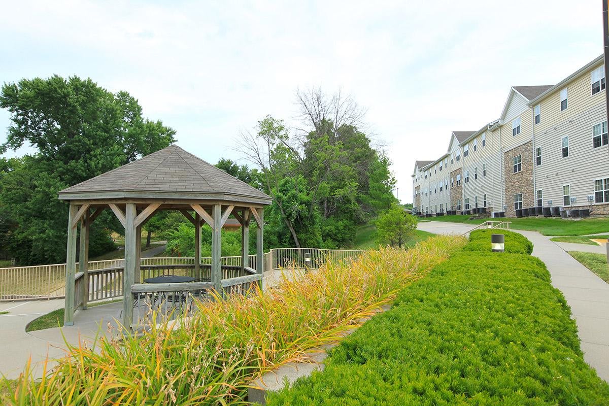 A scenic view featuring a gazebo by a path, surrounded by lush greenery and residential buildings in the background.