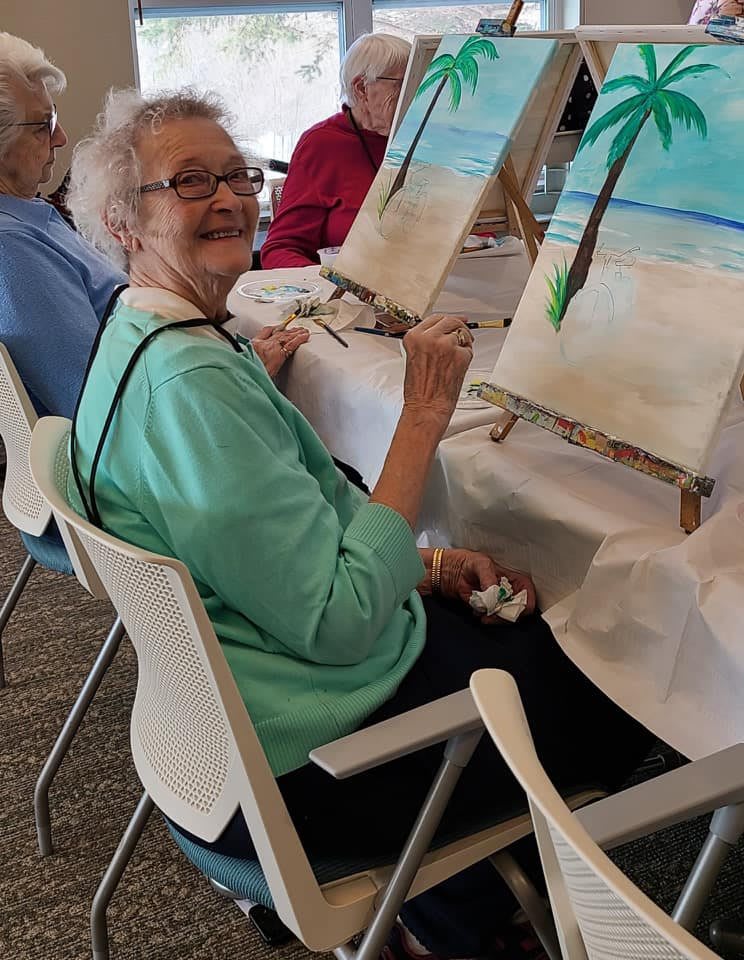 An elderly woman smiling while painting a beach scene on a canvas during an art class.