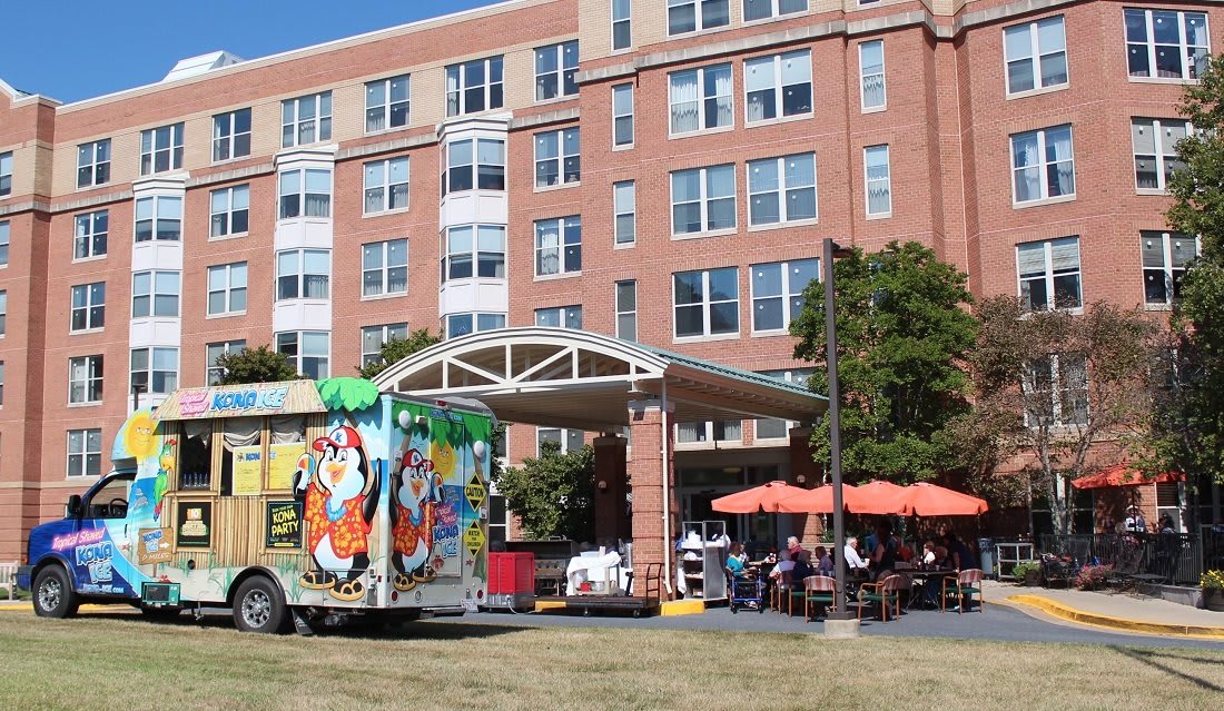 A vibrant food truck parked outside a large building with outdoor seating and several people enjoying the atmosphere.