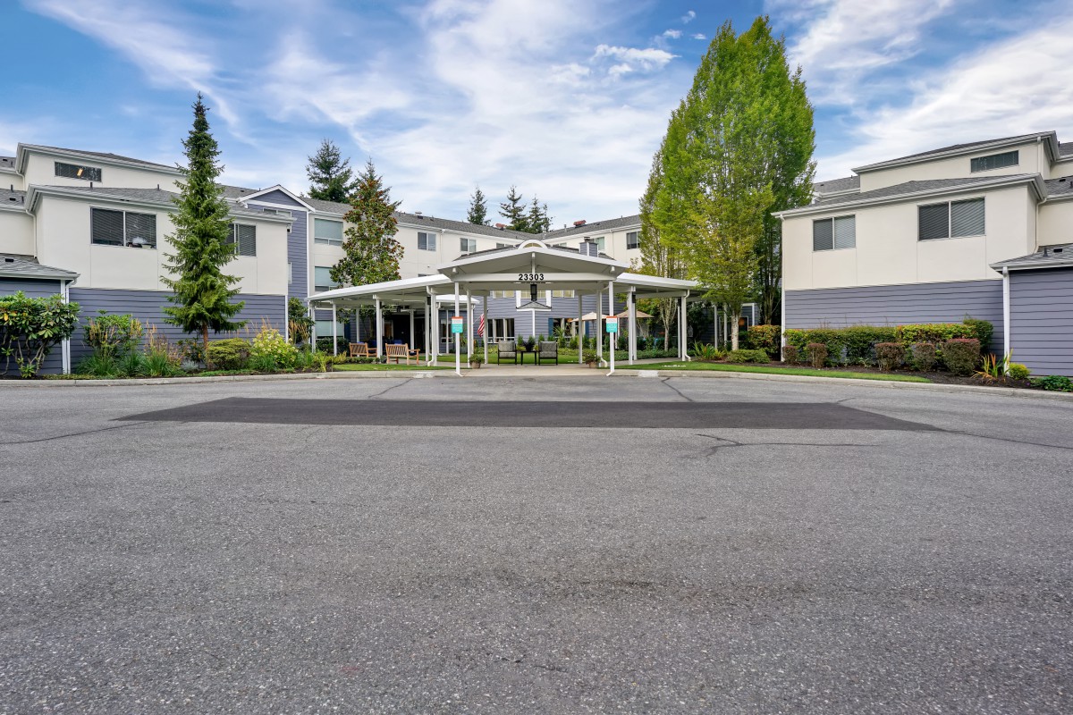 A residential apartment building with a covered entrance, surrounded by trees and a parking lot under a partly cloudy sky.