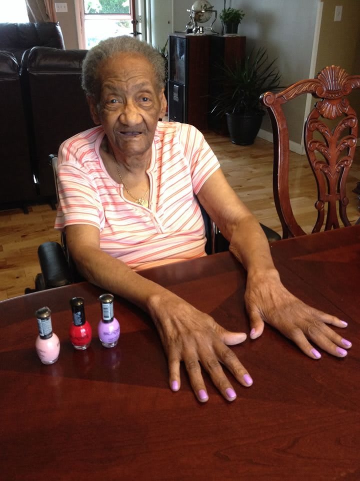 An elderly woman with freshly painted nails sits at a table, smiling.