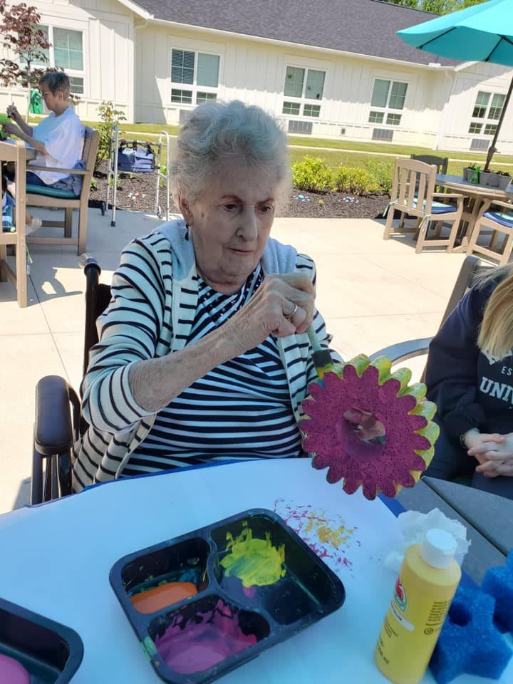 An elderly woman engages in an outdoor arts and crafts activity, painting a colorful item while seated in a wheelchair.