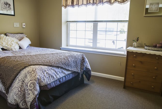 A cozy bedroom featuring a bed with patterned bedding, a large window, and a wooden dresser.