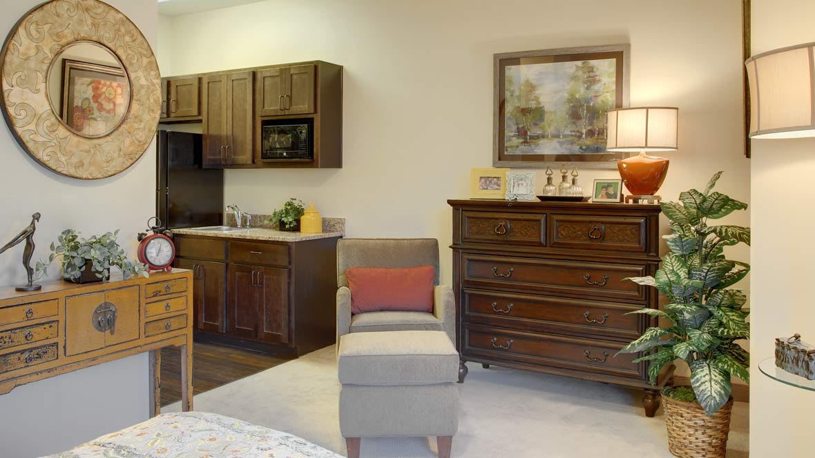 A cozy living area with a kitchenette, featuring a wooden dresser, a comfortable chair, and decorative artwork.