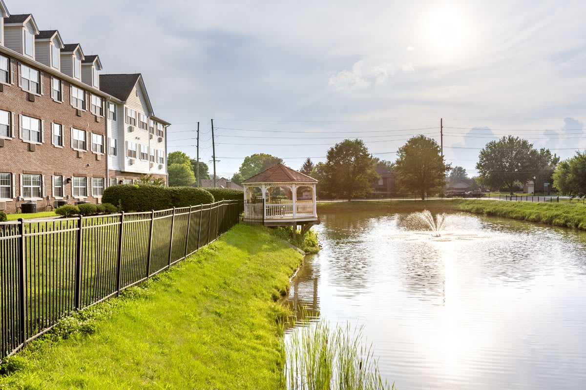A tranquil lakeside view featuring a gazebo, residential building, and lush greenery under a bright sun.